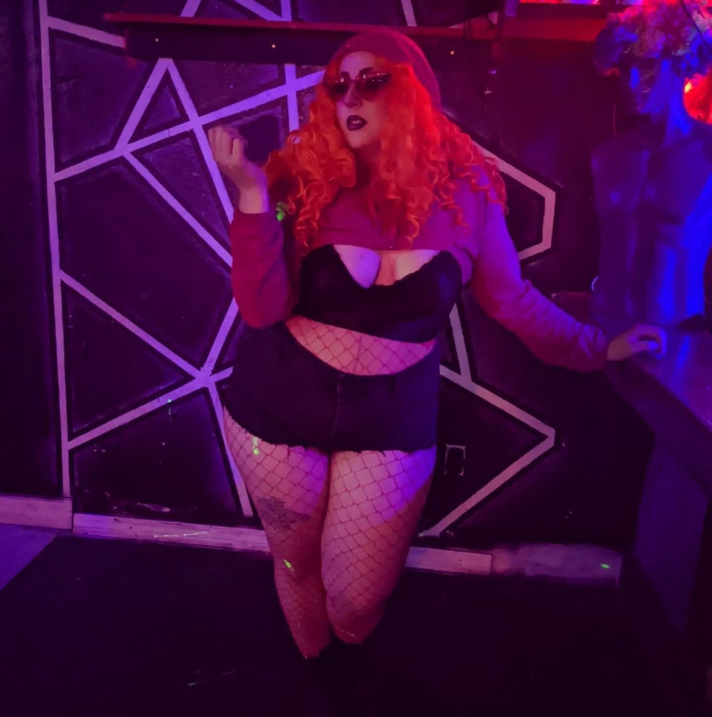 pale person in red curly wig and black outfit in front of geometric pattern
